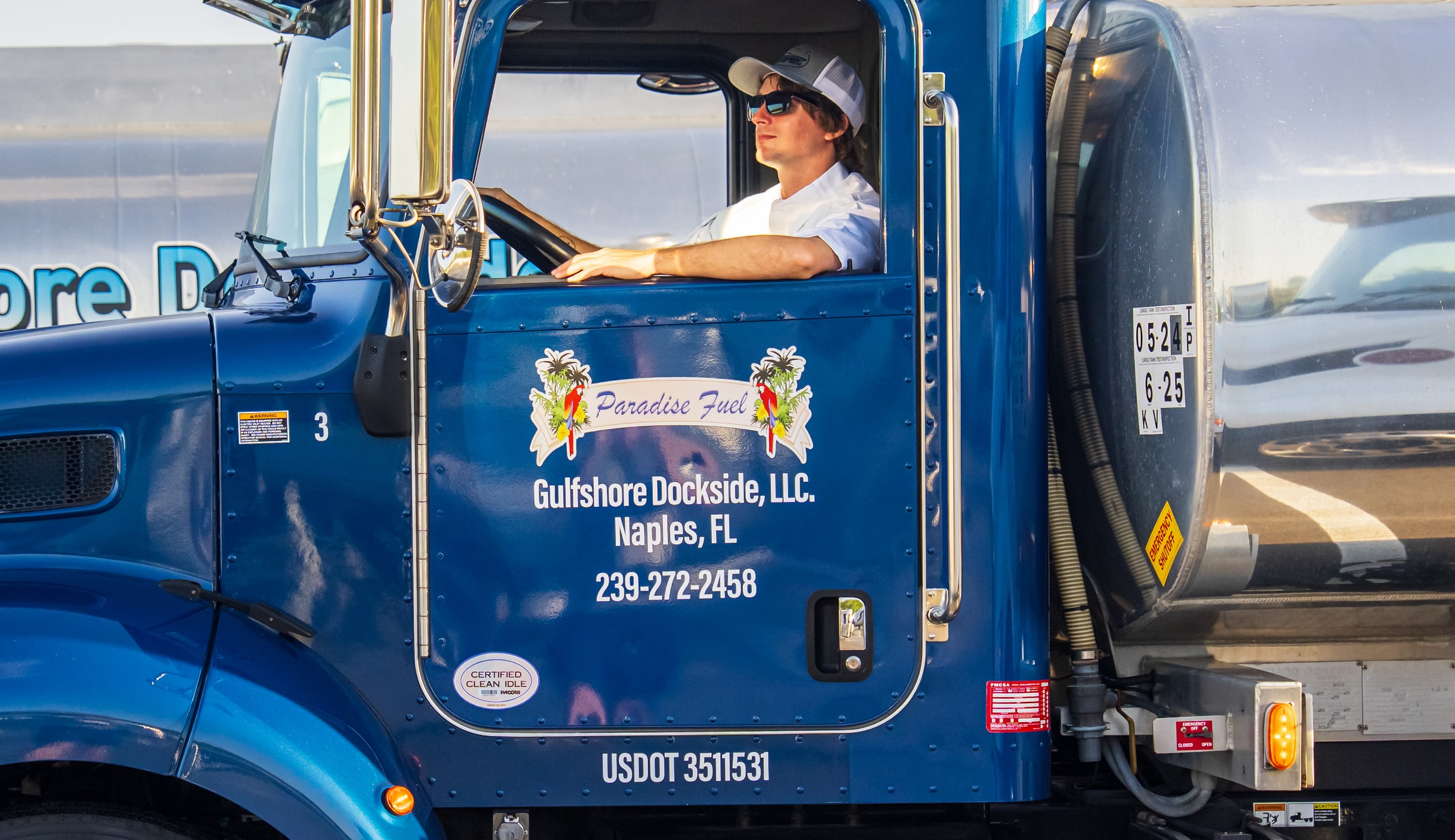 The door of tanker number three: the Paradise Fuel ribbon with its parrots and palms, and lettered directly beneath it, Gulfshore Dockside, LLC., Naples, FL, 239-272-2458. The driver sits at the wheel with his arm out of the window.